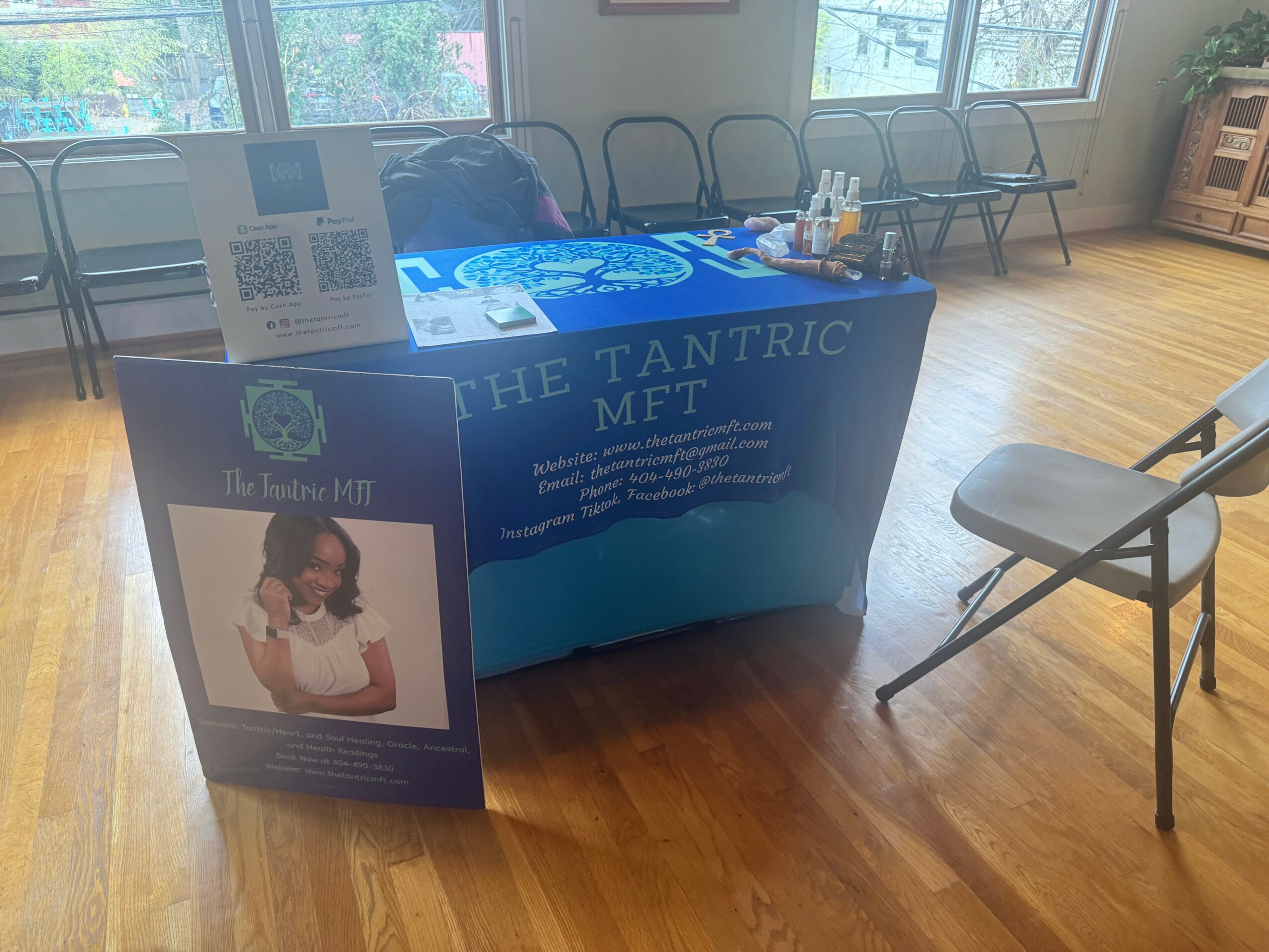 Table with blue tablecloth displaying promotional materials, a poster with a child, and chairs in a room with wooden floor and windows.