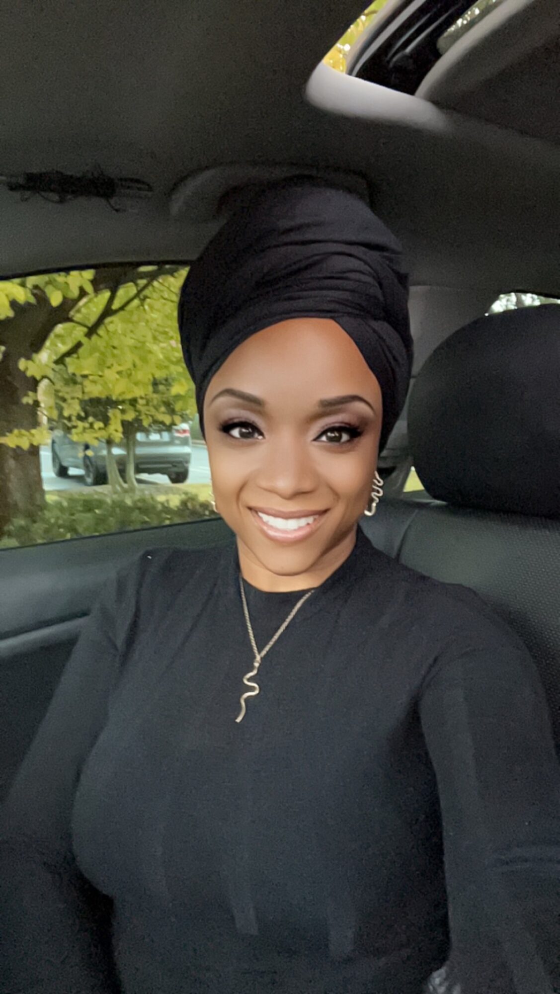 Woman with a headwrap smiling in car, trees visible outside window.