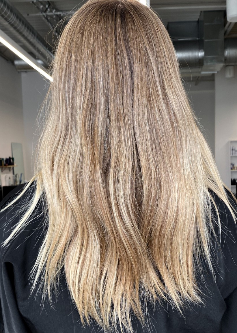 Back of a person with long, wavy blonde hair, wearing a dark top, in an indoor setting.