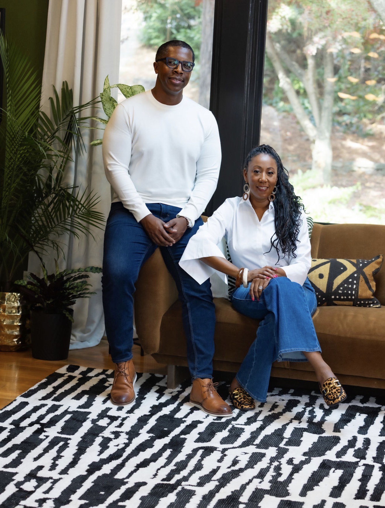 Two women pose indoors, one standing and one sitting on a sofa, with a window and trees outside.