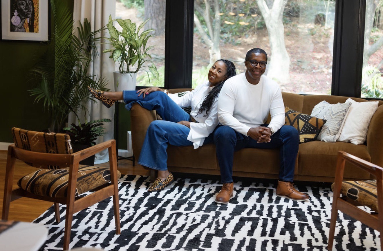 Two people sit on a sofa in a living room with large windows, plants, and a black-and-white patterned rug.