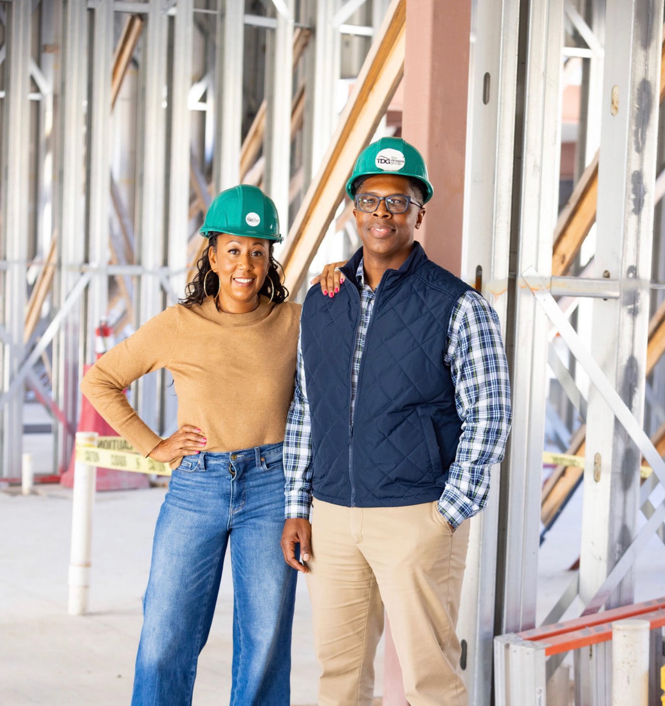 Two people wearing green hard hats standing in front of construction framework, smiling at the camera.