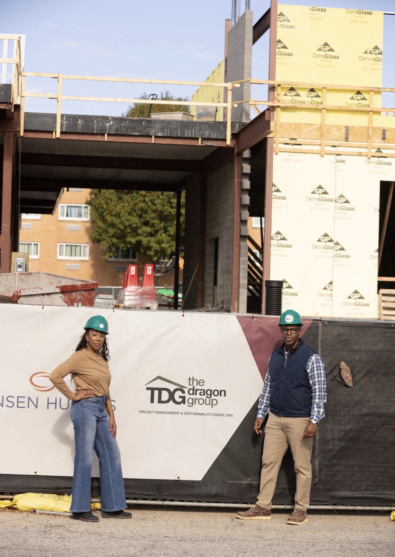 Two construction workers wearing helmets stand in front of a construction site with a banner displaying logos.