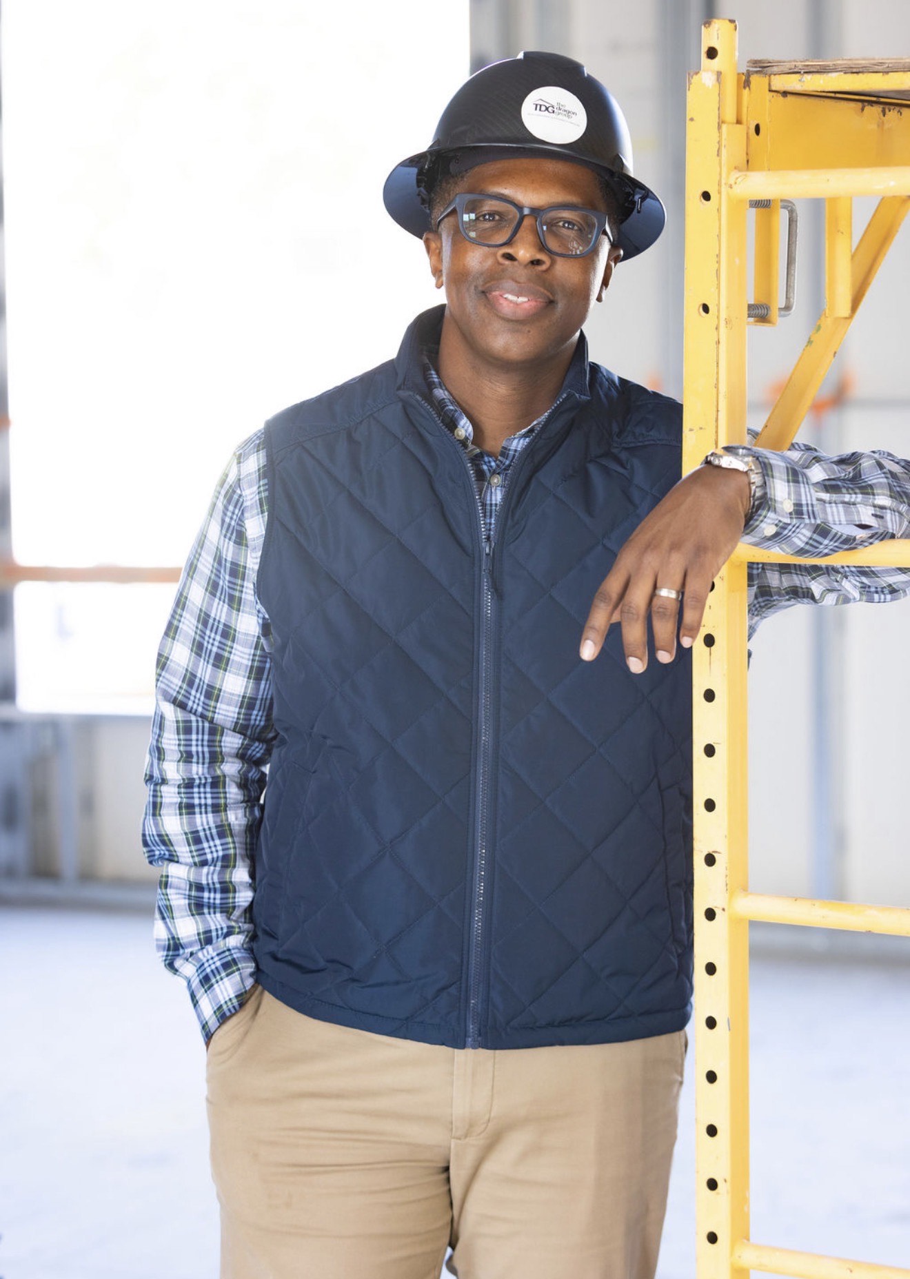 Man in safety helmet and glasses leaning on yellow scaffolding in industrial setting.