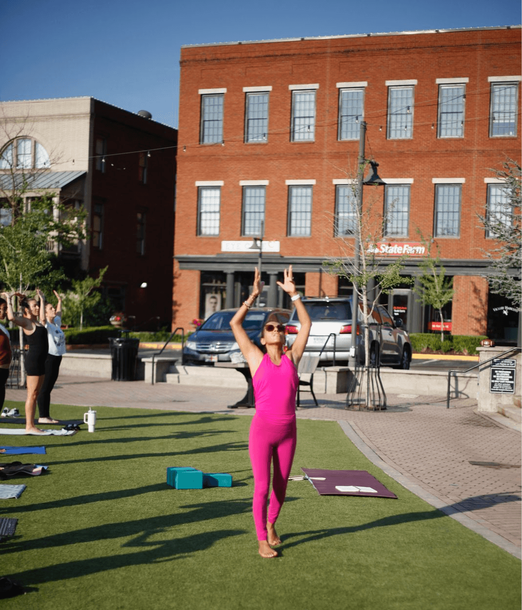 Woman in pink workout clothes standing on grass, raising arms, with buildings and parked cars in background.