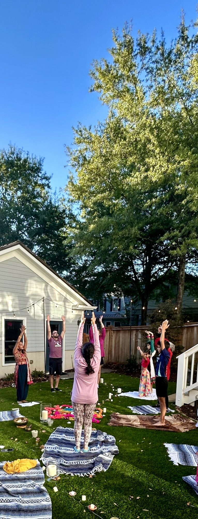 People practicing yoga outdoors on mats in a grassy yard with trees and a house in the background.
