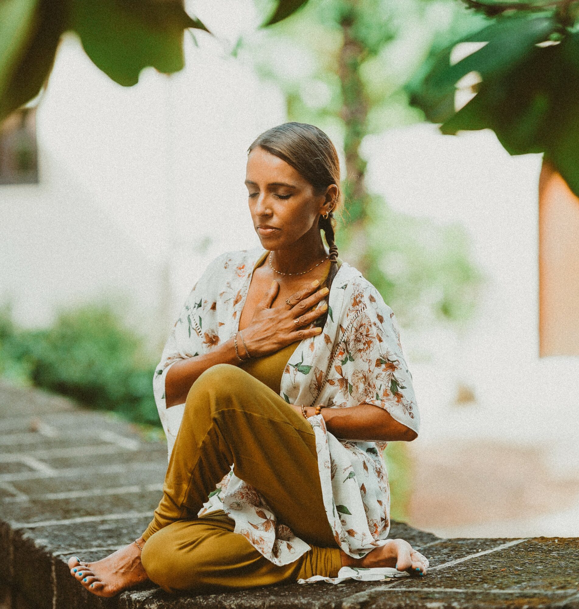Woman sitting cross-legged outdoors with hand on chest, eyes closed, surrounded by greenery.