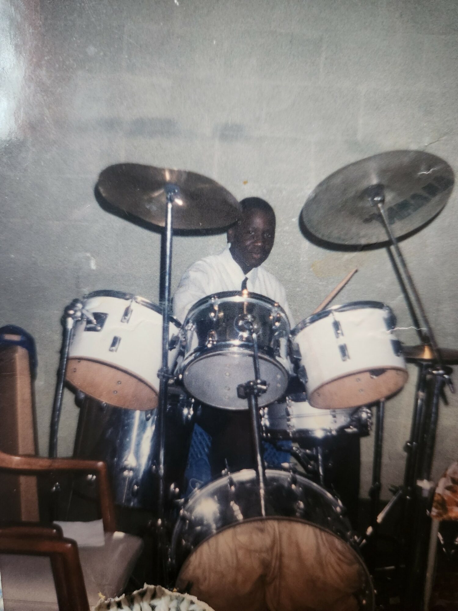 Person standing behind a drum set with cymbals in a room with plain wall background.