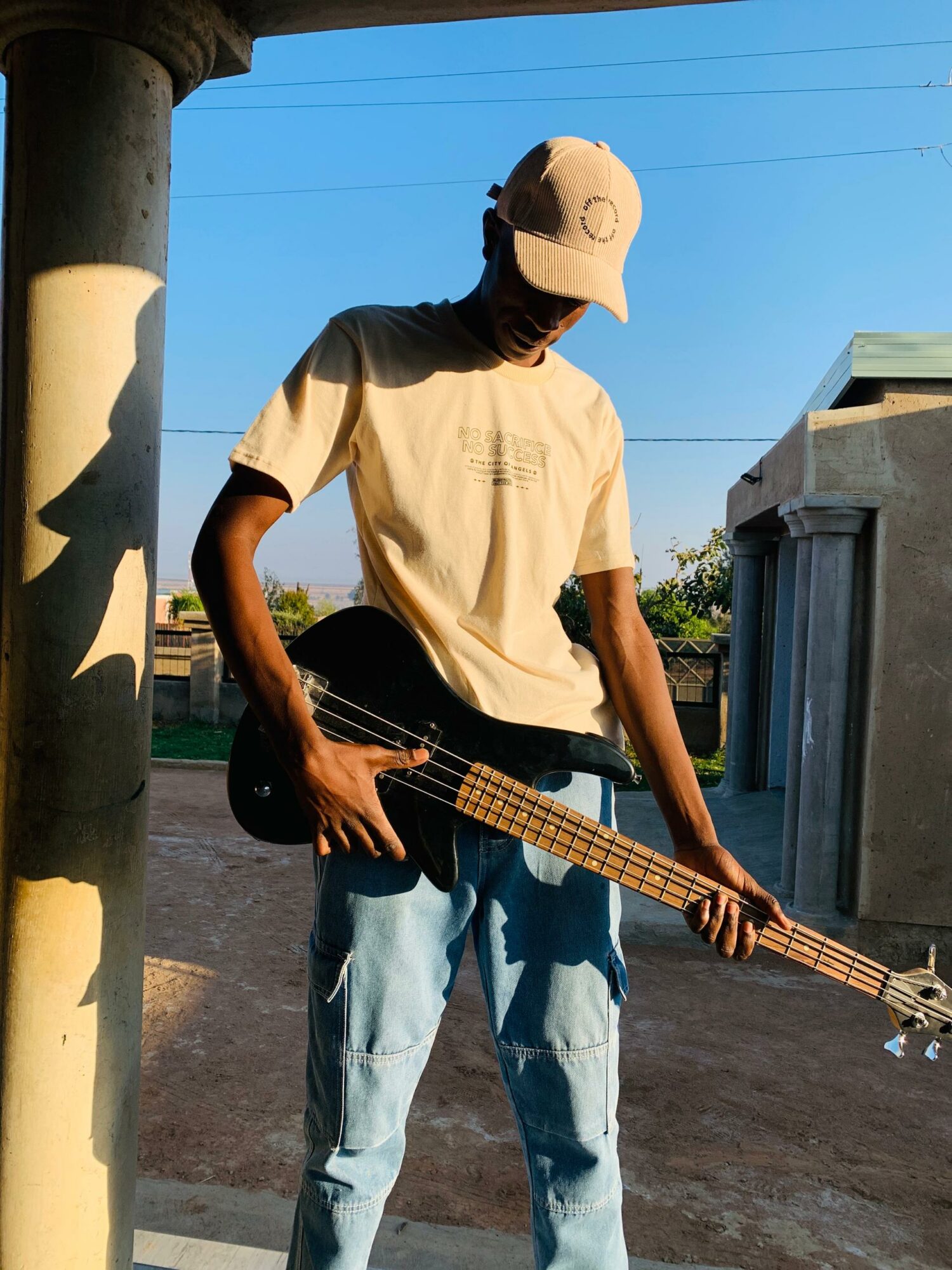 Person wearing a cap and t-shirt playing a bass guitar outdoors under a clear blue sky.