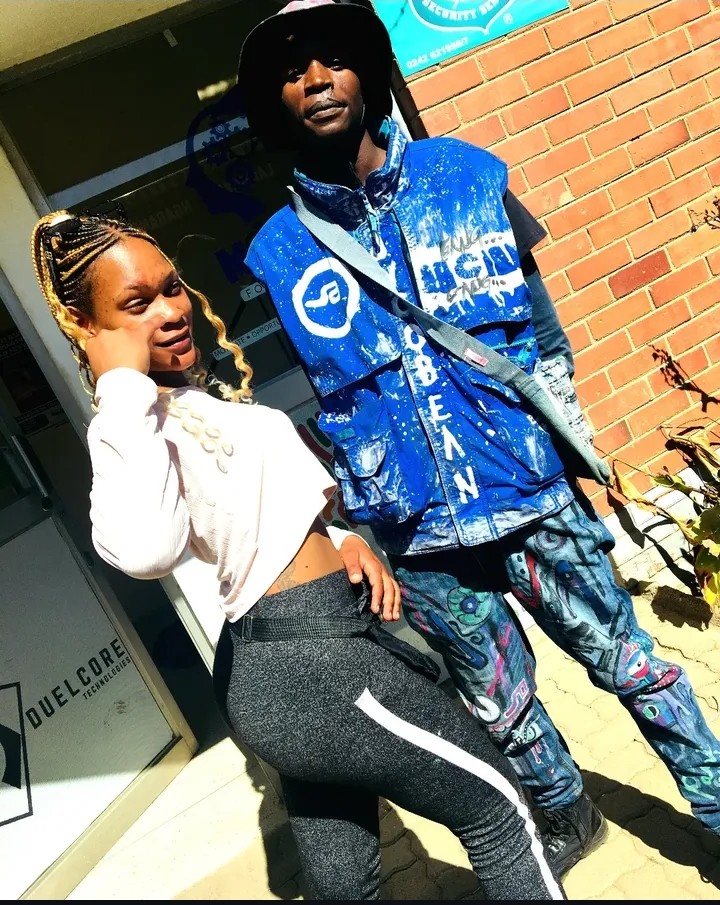 Two people standing outside a building with a brick wall, one woman and one man, both dressed casually.