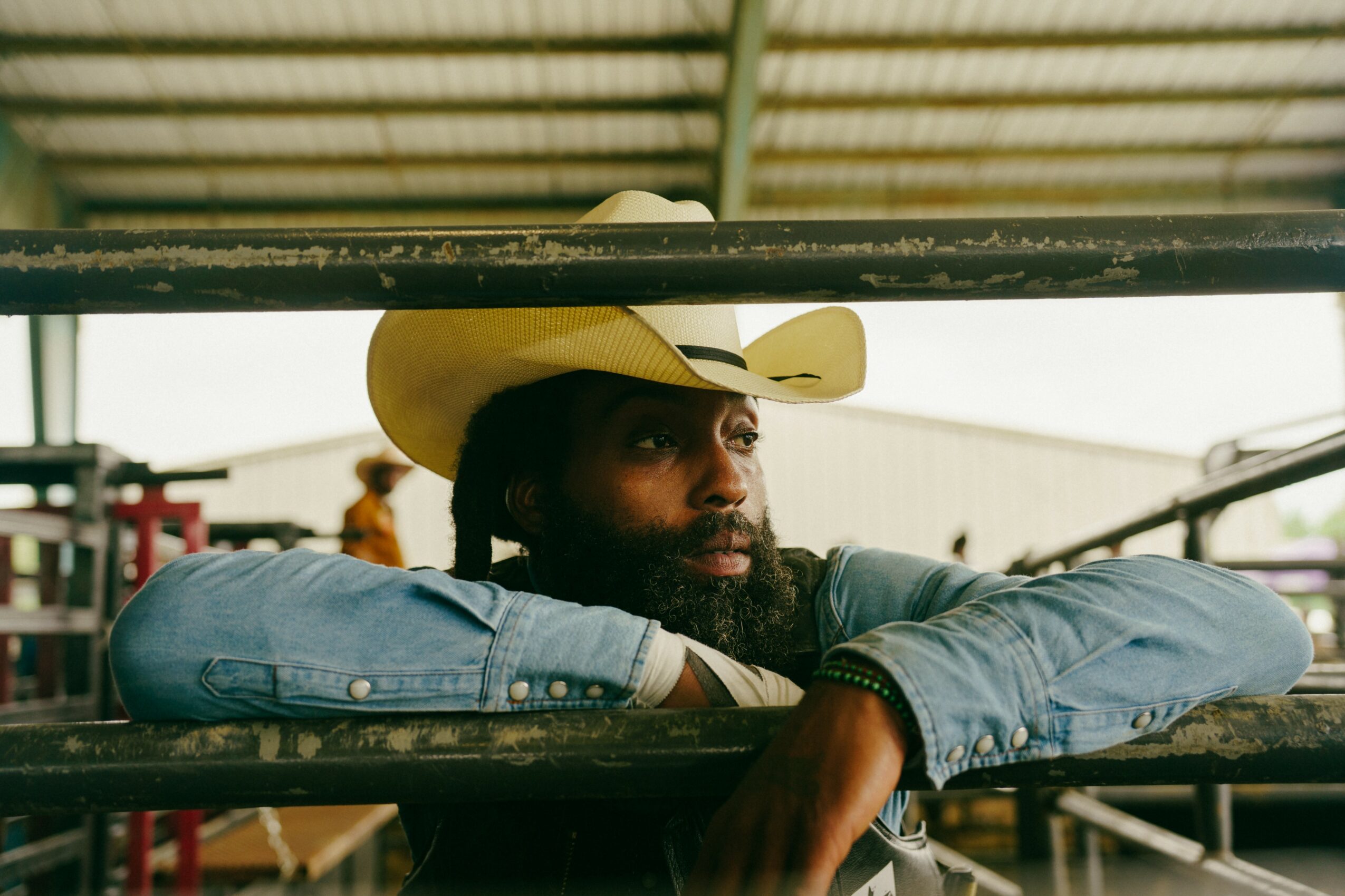 Man wearing a cowboy hat resting his arms on a metal railing, looking into the distance.