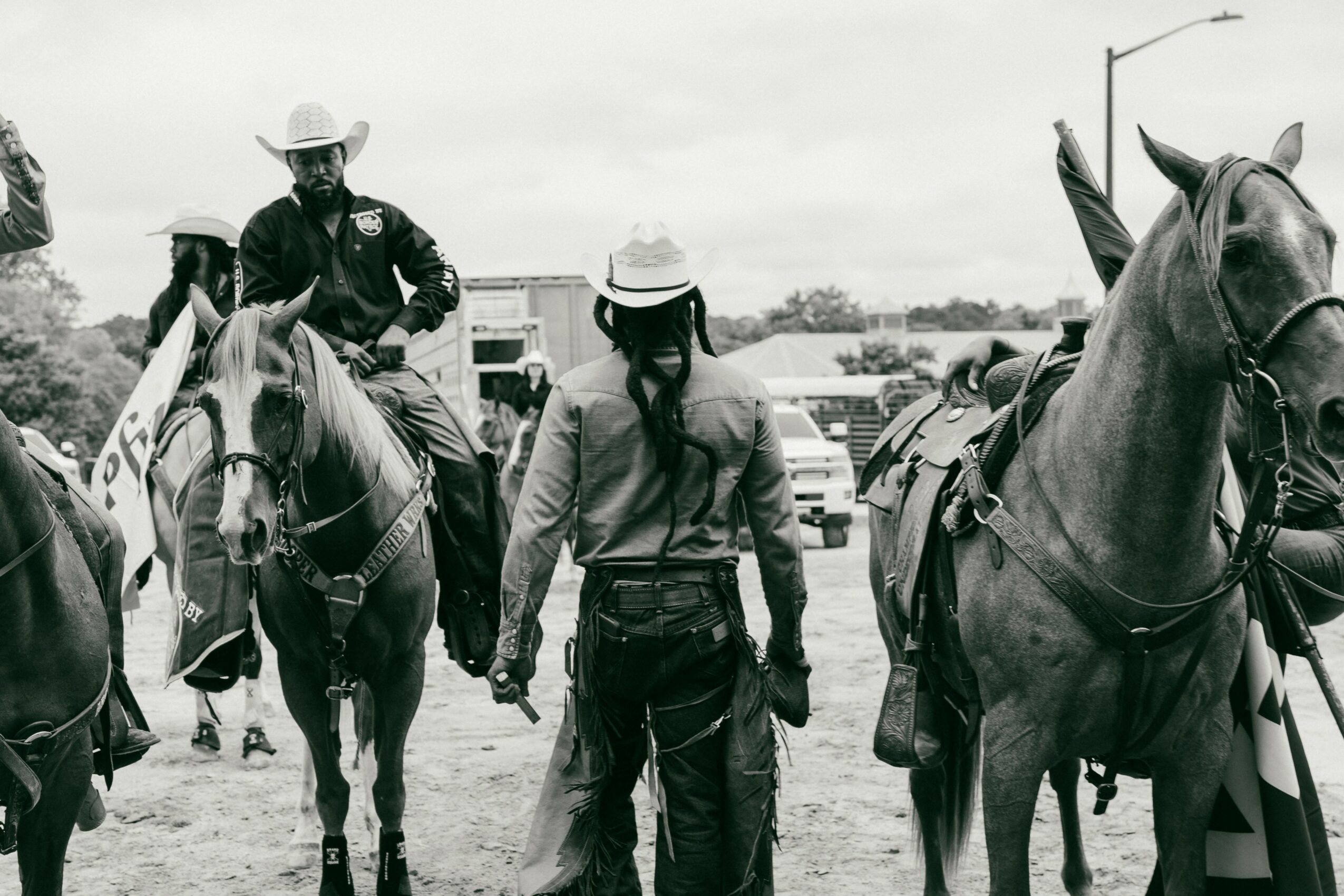 Two people on horseback and one person standing with horses outdoors, with vehicles and trees in background.