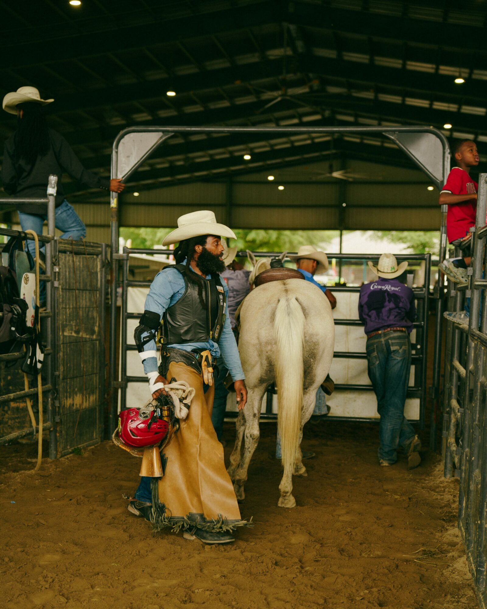 Person wearing cowboy hat and face mask standing near a white horse inside a covered area with other people around.