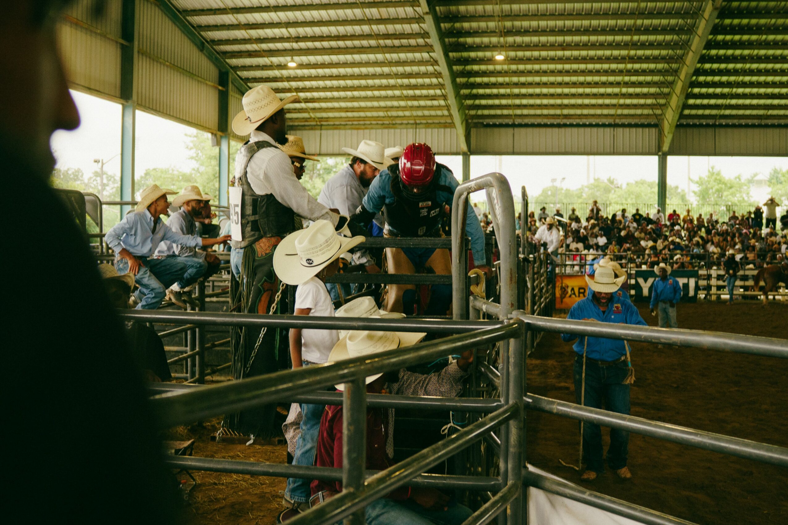 People gathered in a covered arena, some with hats and guitars, watching a rodeo event with a crowd in the background.