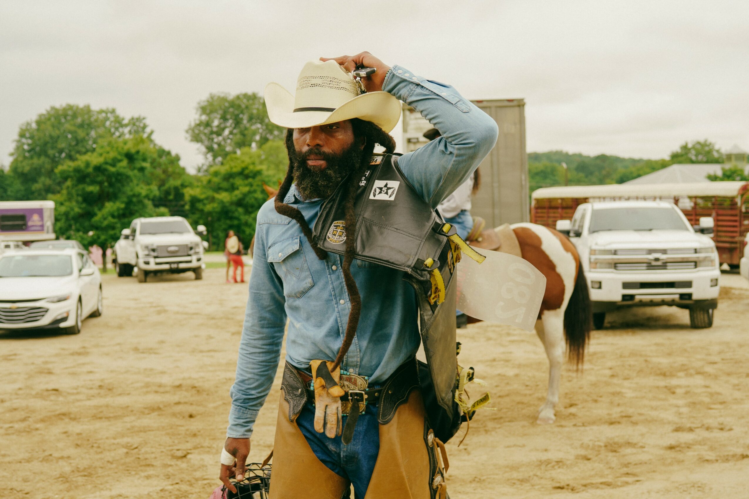 Person wearing a cowboy hat and denim shirt standing outdoors with horses and parked cars in background.