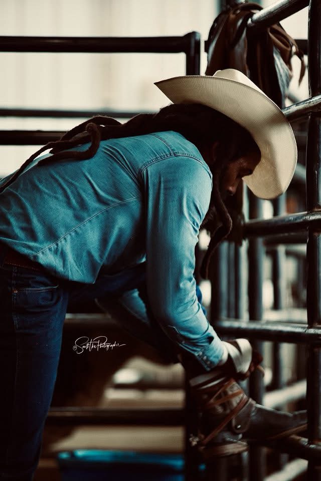 Person wearing a large cowboy hat and blue shirt, leaning forward, adjusting a saddle on a horse in a stable.