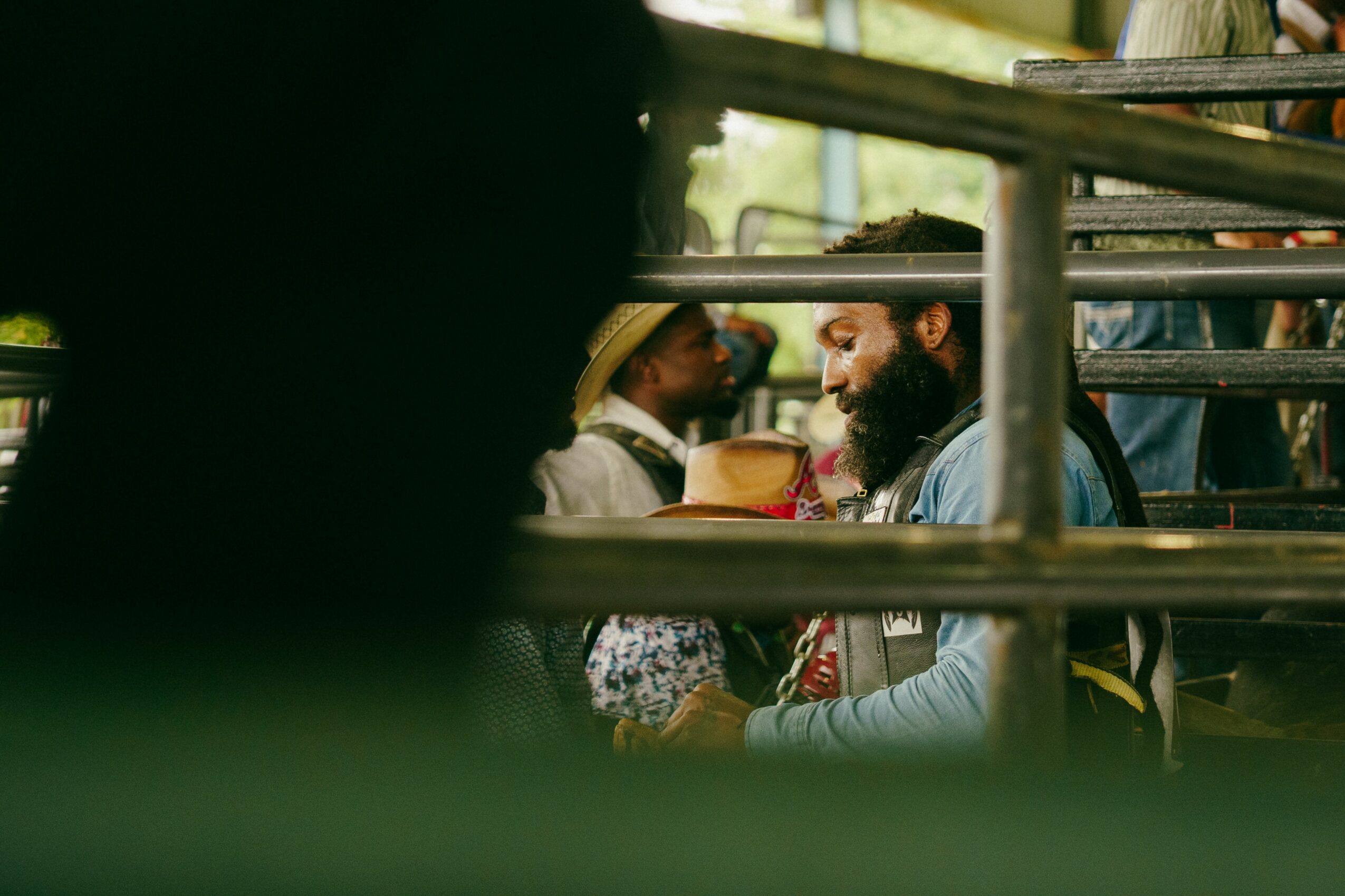 Two men sitting on a bench, one with a beard and hat, engaged in conversation, with blurred foreground elements.