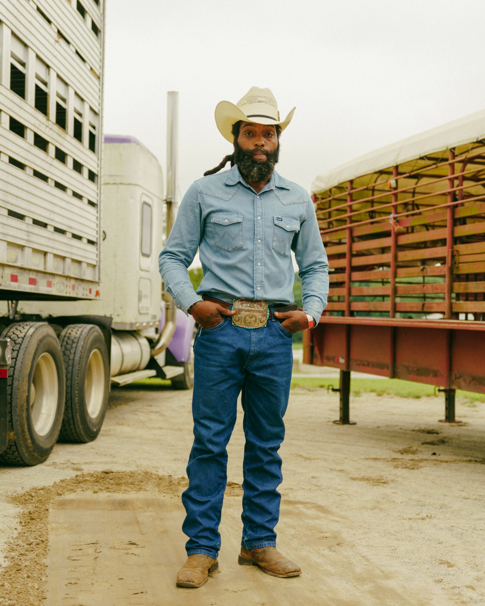Man in cowboy hat, denim shirt, jeans, and boots standing outdoors near trucks and a trailer.