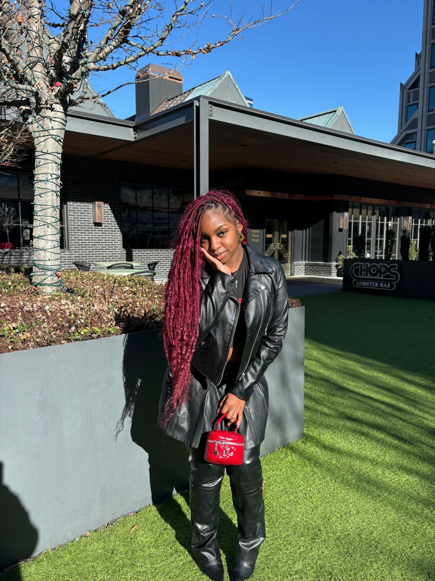 Woman with long braided hair wearing black leather jacket and pants, holding a red purse, standing outdoors on grass.