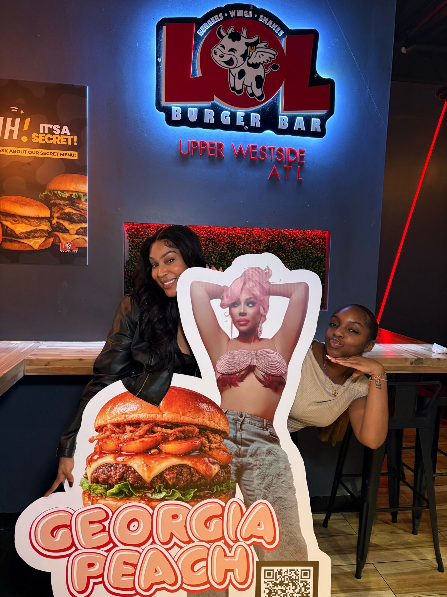 Two women sitting at a table in front of a large cutout of a woman with pink hair, inside a burger restaurant.