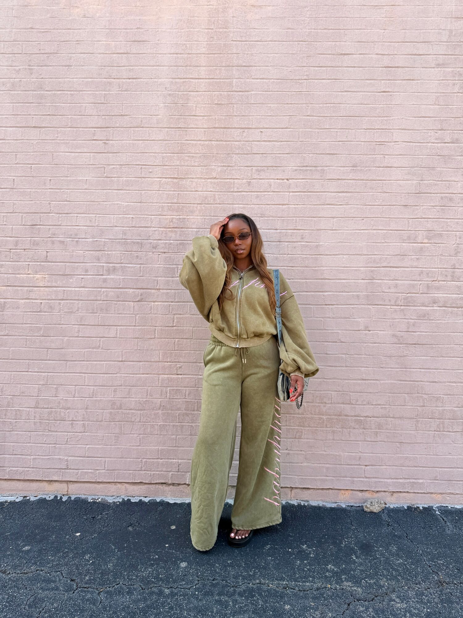 Person standing against a pink brick wall, wearing a beige outfit and holding a bag.