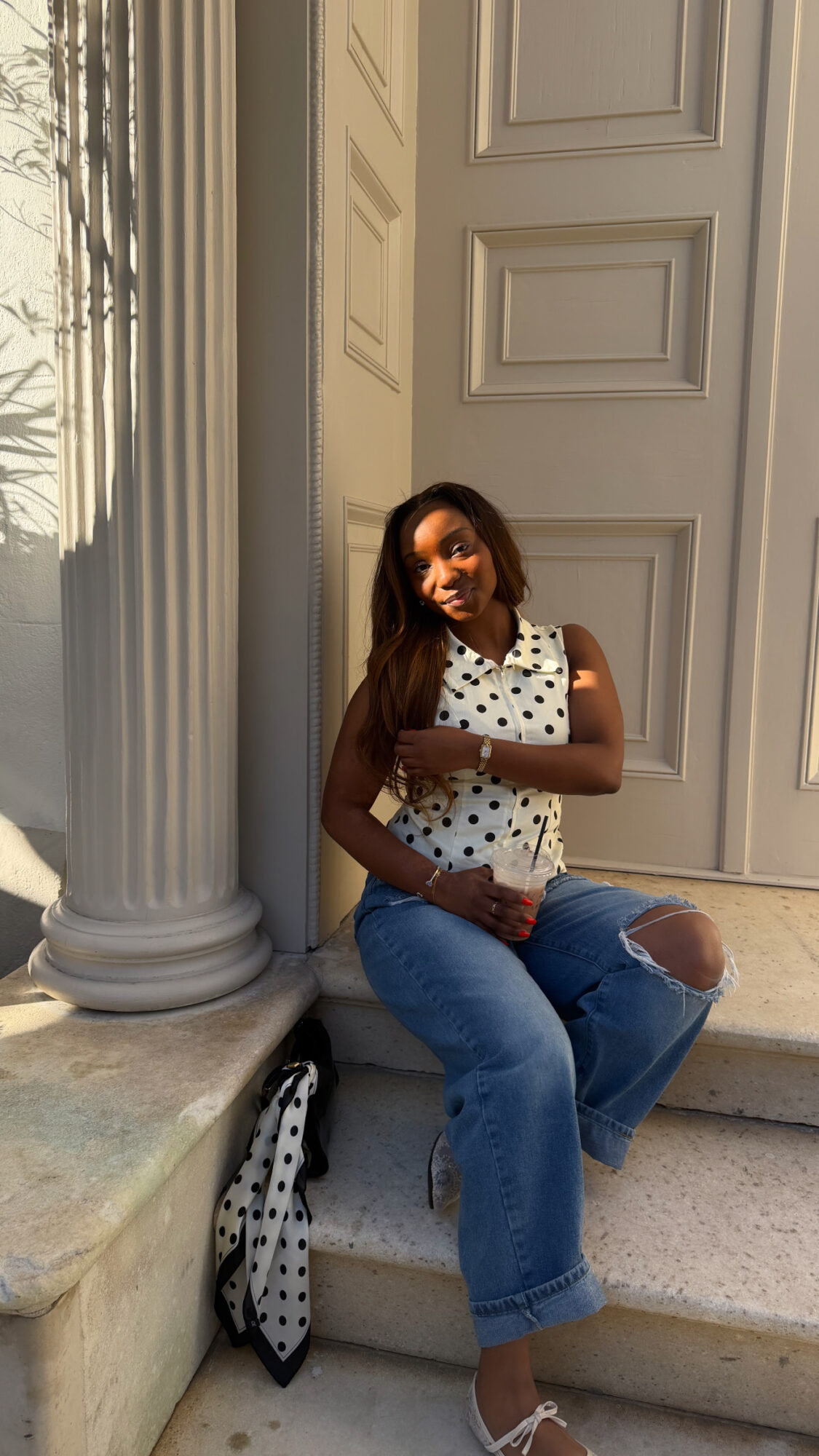 Woman sitting on stairs next to a large column and a door, smiling, wearing a polka dot top and ripped jeans.