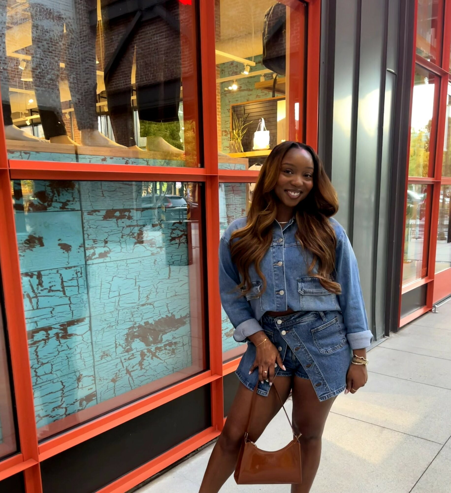 Smiling woman with long hair in denim jacket and shorts, holding a brown bag, standing outside a building with large windows.