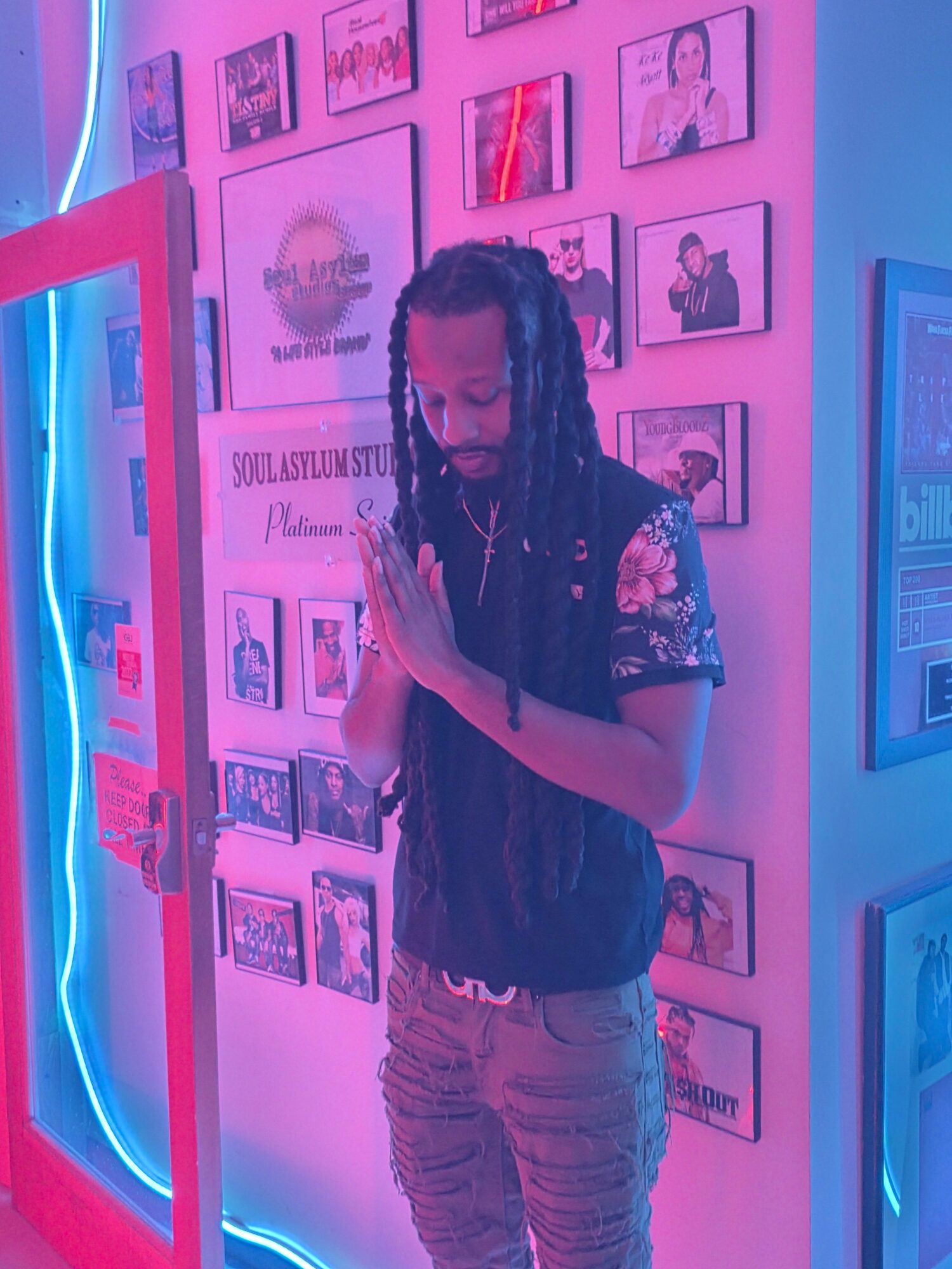 Person with long dreadlocks and floral shirt standing with hands together in front of a wall with framed pictures, illuminated by pink and blue lights.