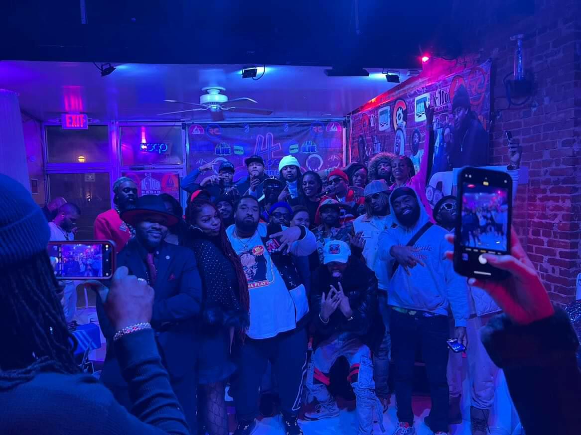 Group of people gathered in a dimly lit indoor space with colorful lighting, some taking photos with smartphones.