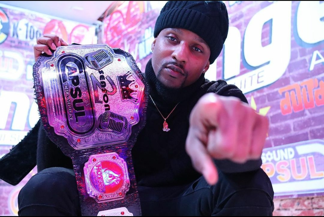 Man in black beanie holding a pink wrestling championship belt, pointing at the camera, with colorful background banners.