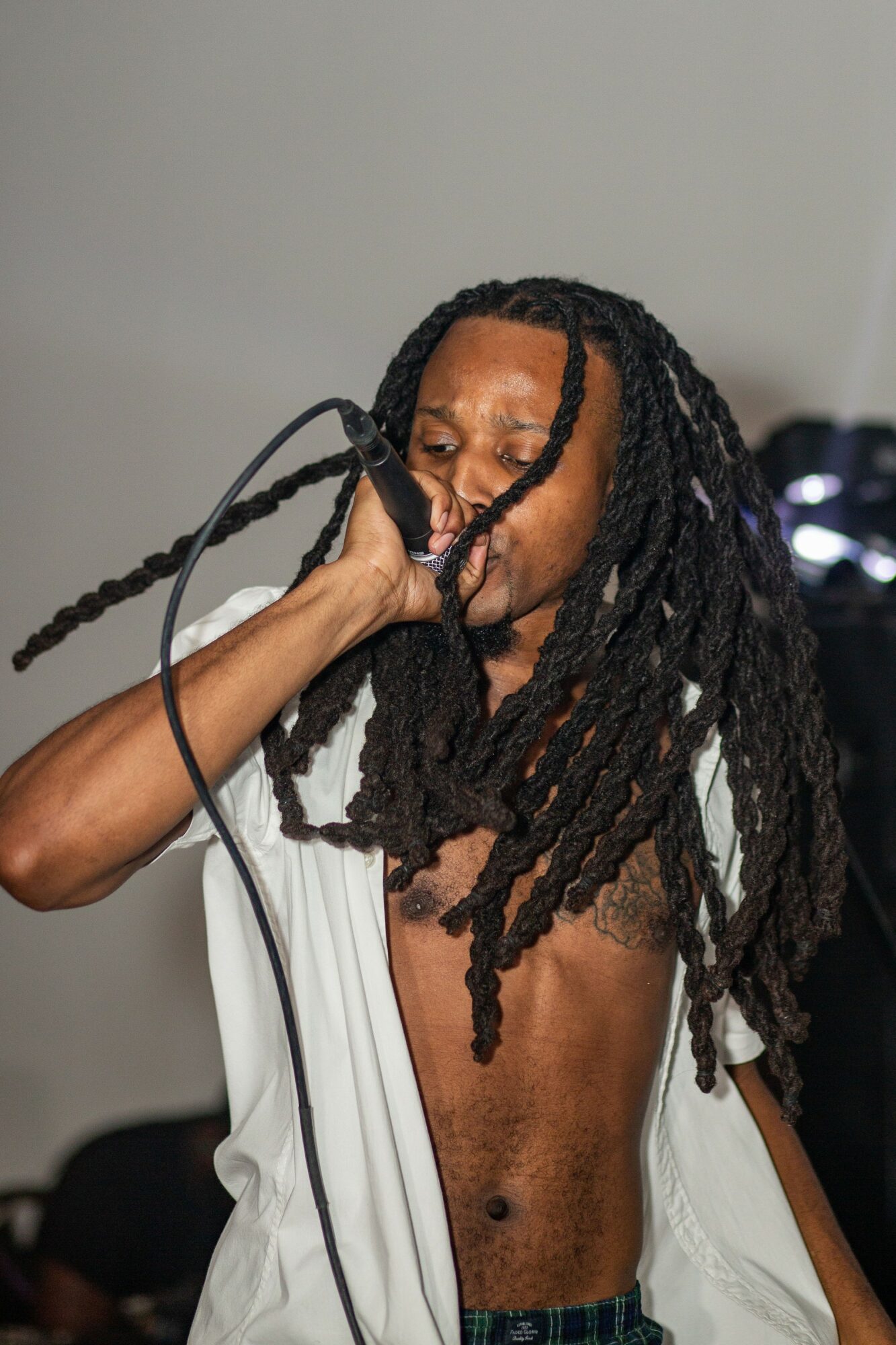 Person with long dreadlocks singing into a microphone, eyes closed, shirt off, in a performance setting.