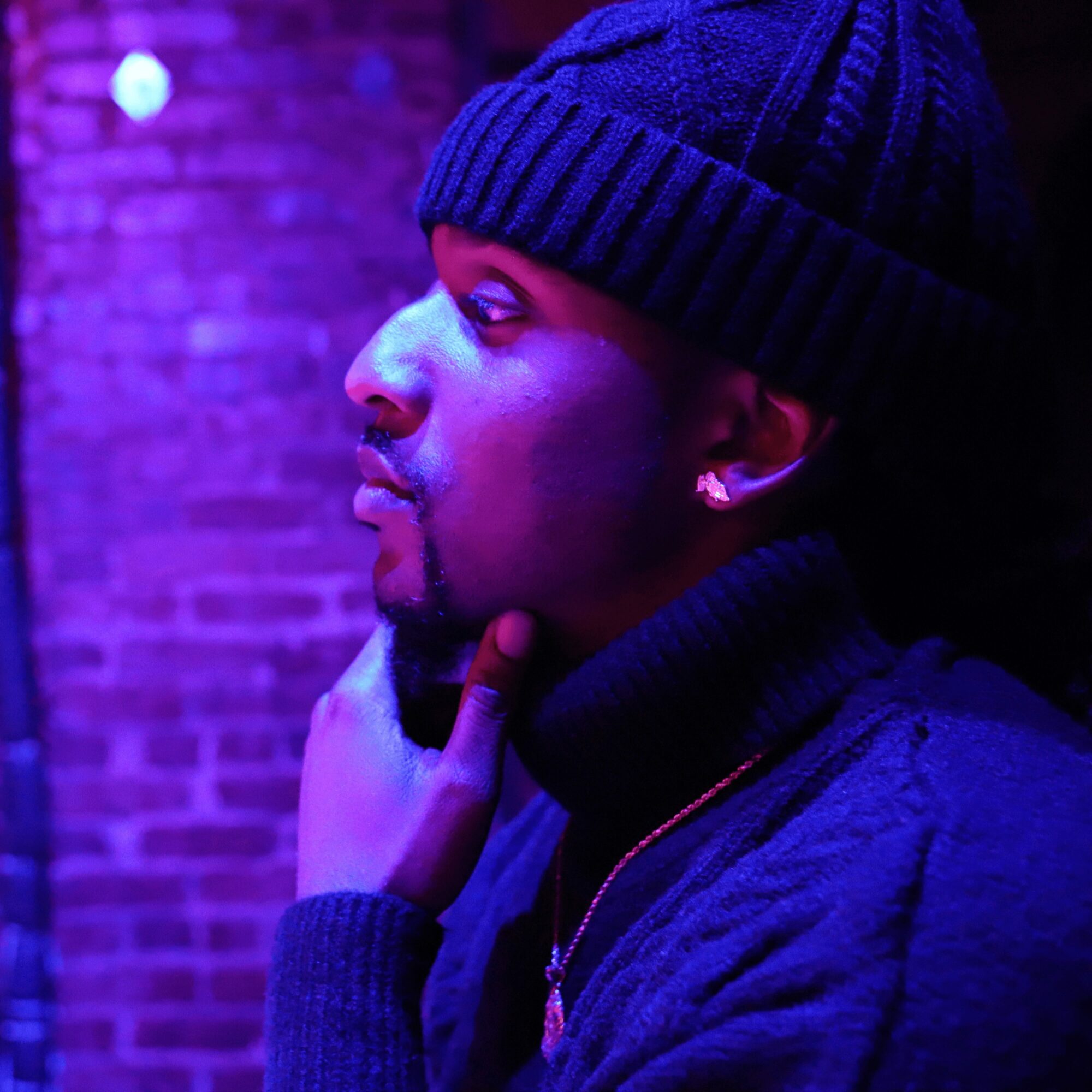 Side profile of a person wearing a beanie and jacket, touching chin, illuminated by purple and blue lighting.