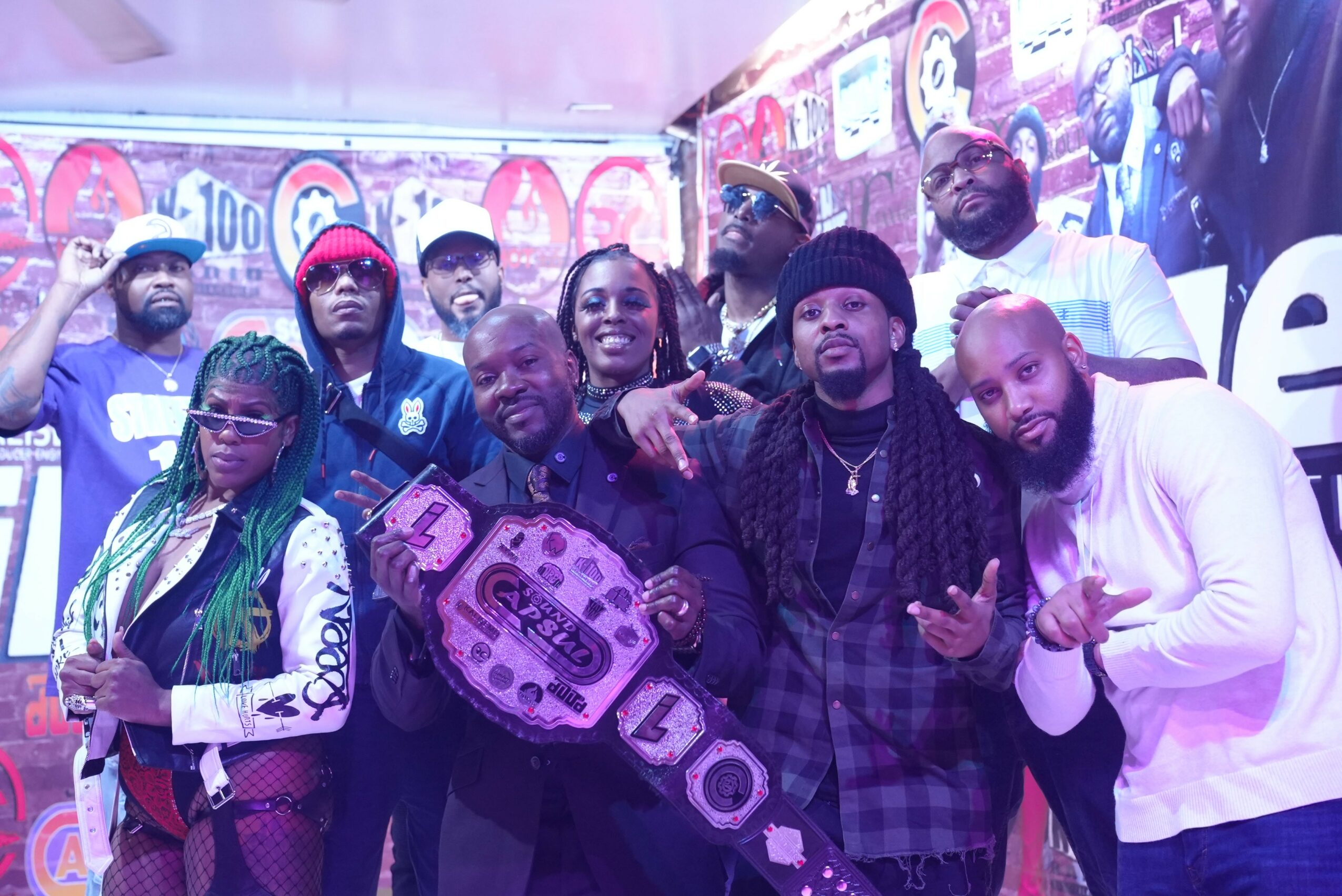 Group of people posing together, some holding a large purple boombox, in front of a colorful backdrop with graffiti-style art.