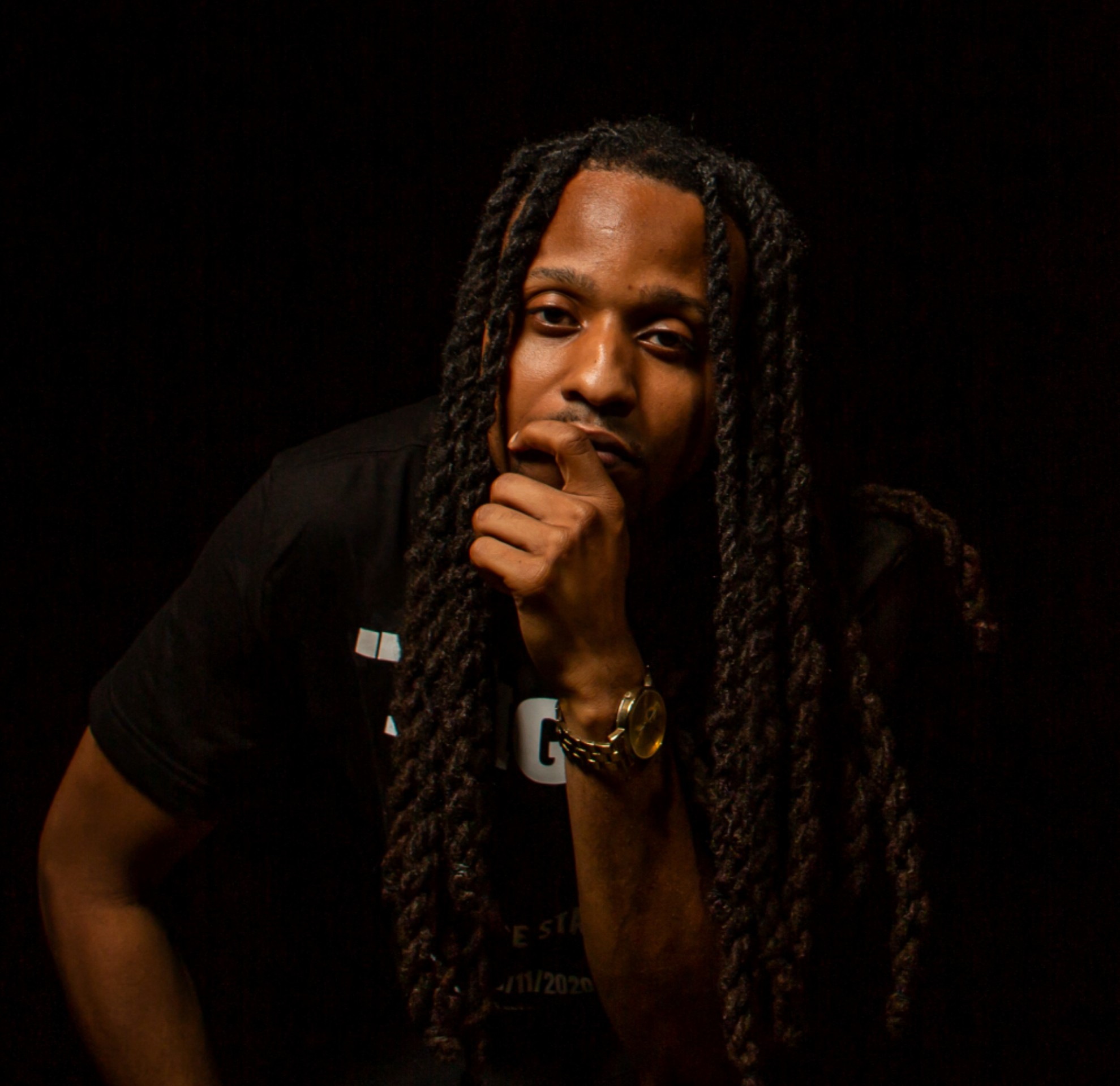 Person with long braided hair resting chin on hand, dark background, wearing dark clothing and a watch.