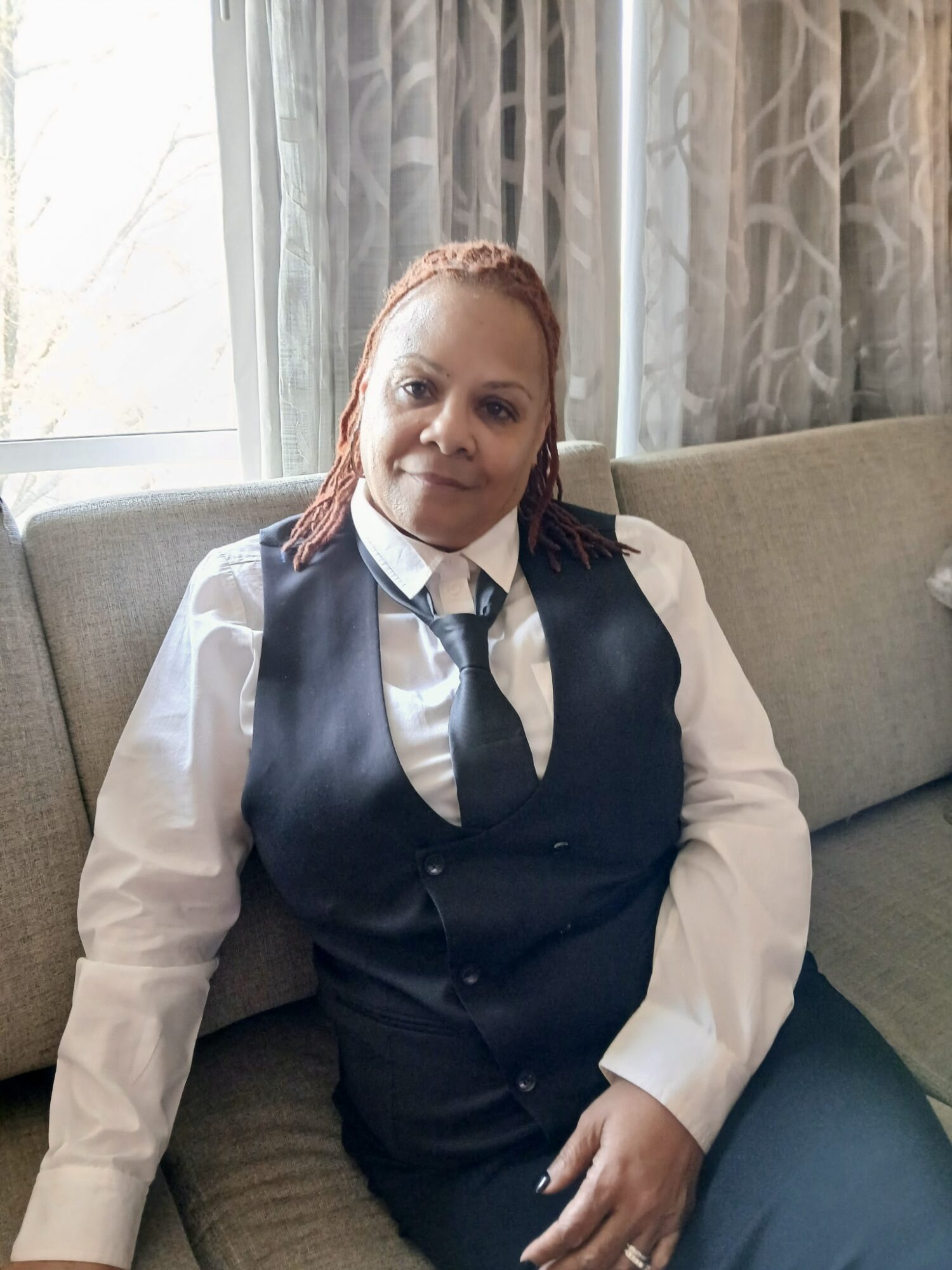 Woman with reddish hair sitting on a sofa, wearing a white shirt, dark vest, and tie, in front of curtains.