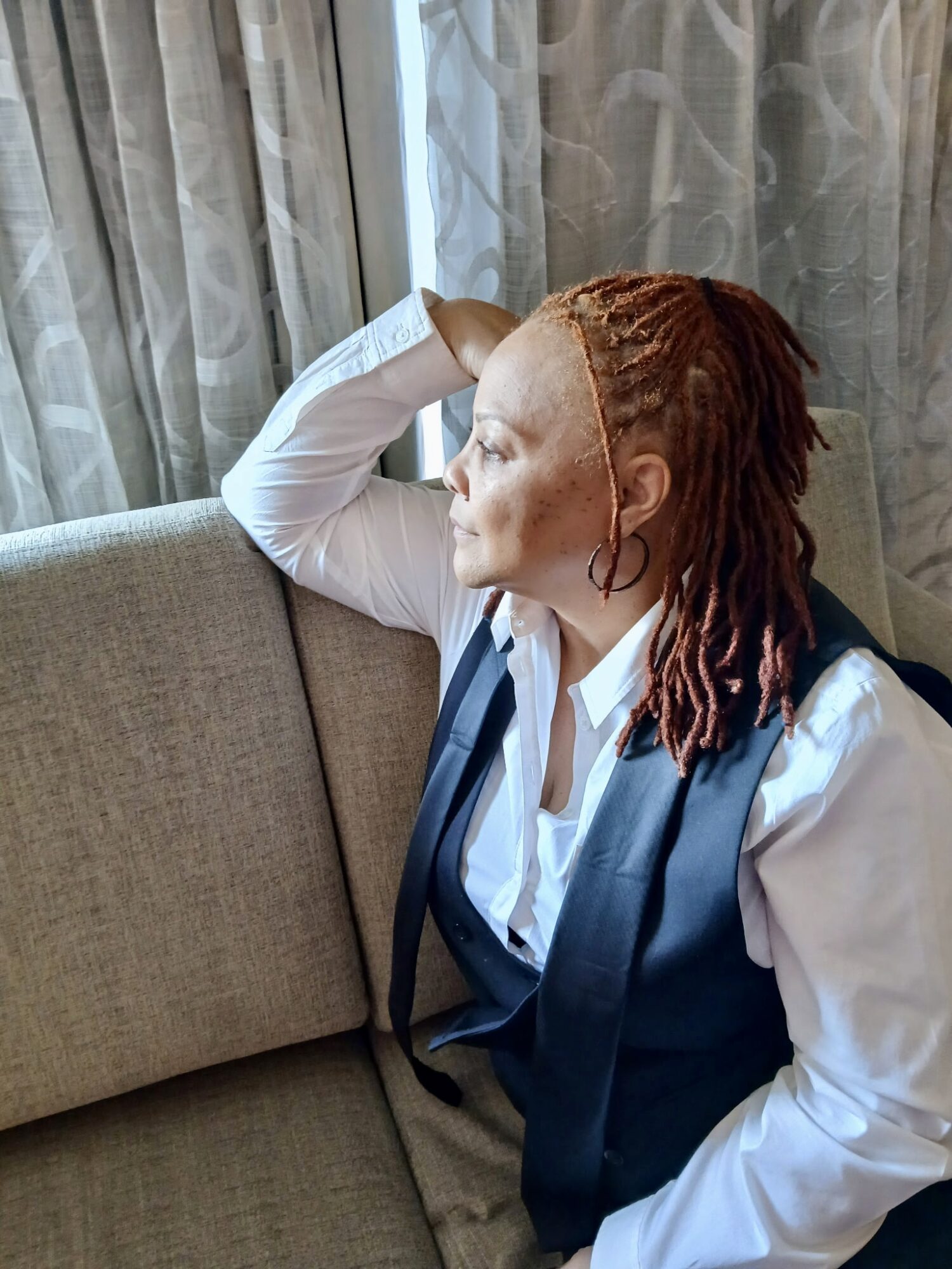 Woman with red dreadlocks sitting on a sofa, resting her arm on the back, looking to the side near curtains.
