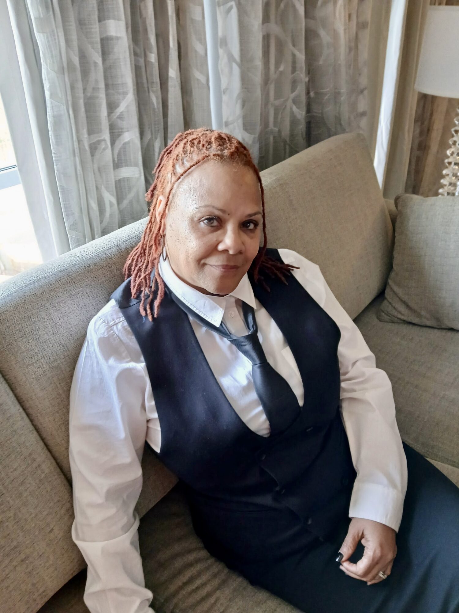Woman with reddish dreadlocks sitting on a sofa in a room with curtains and a lamp.