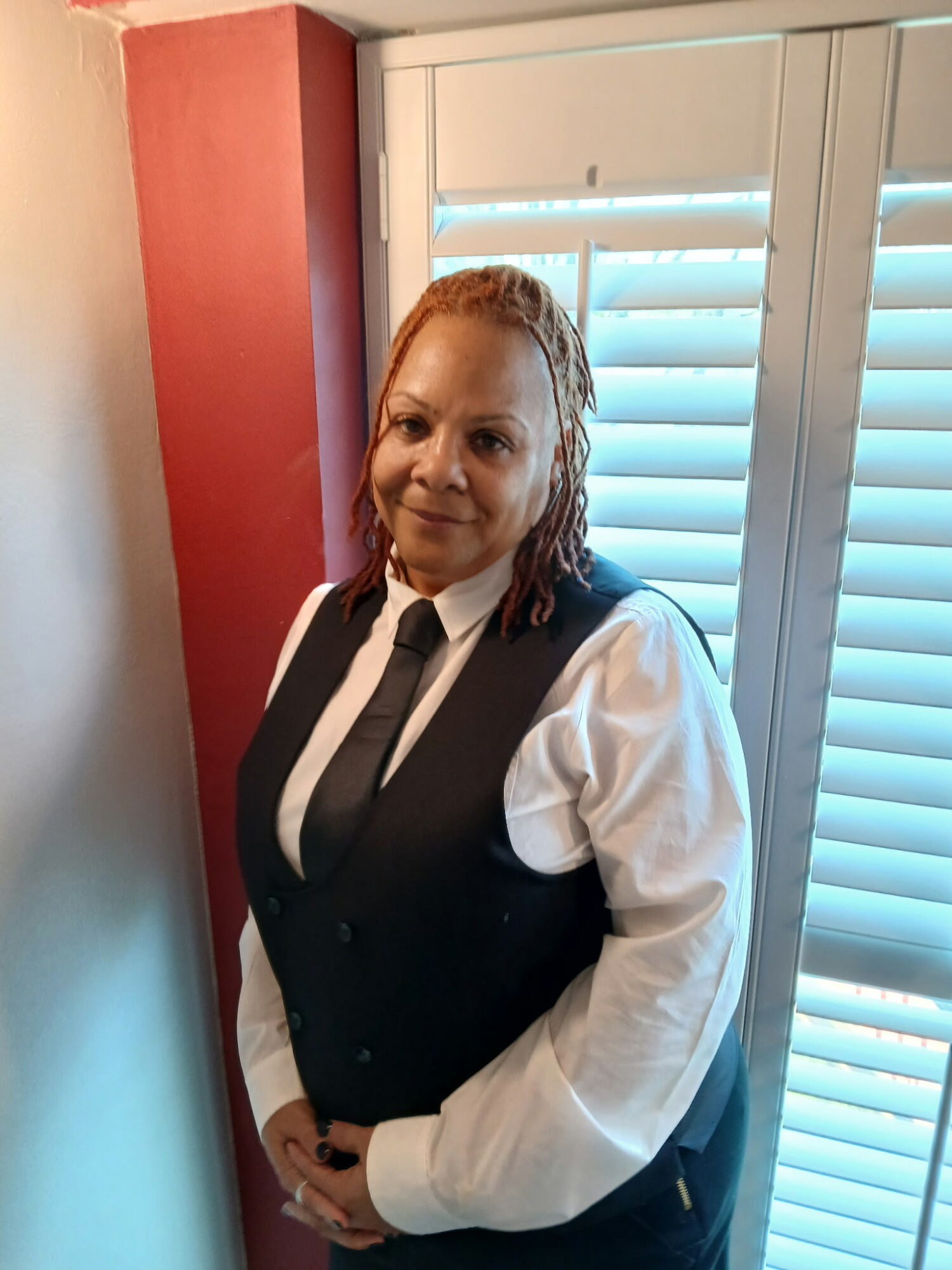 Woman with curly hair in a white shirt and black vest standing indoors near window with shutters.
