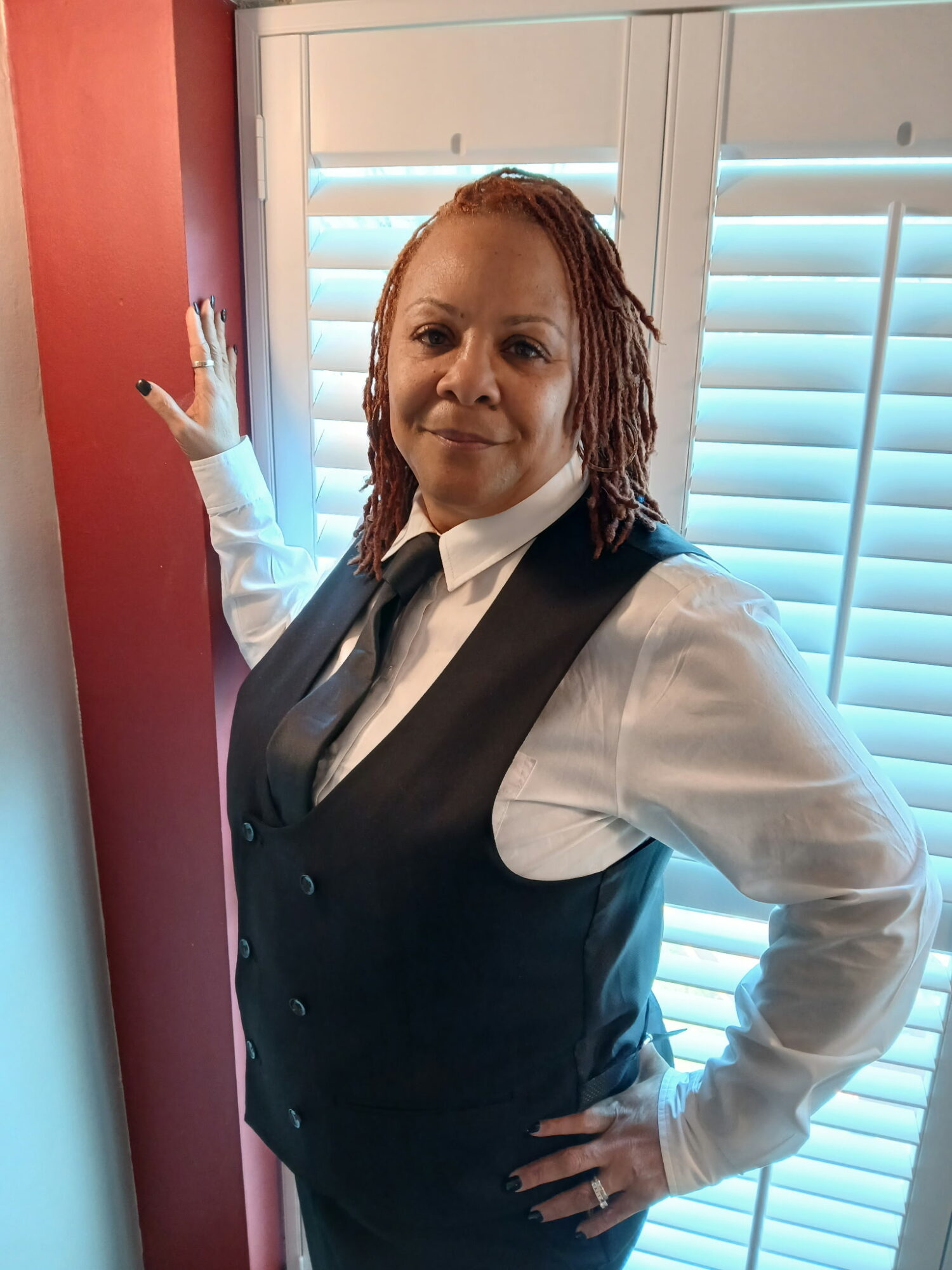Woman with dreadlocks in a vest and white shirt standing near window with blinds, hand on hip, smiling.