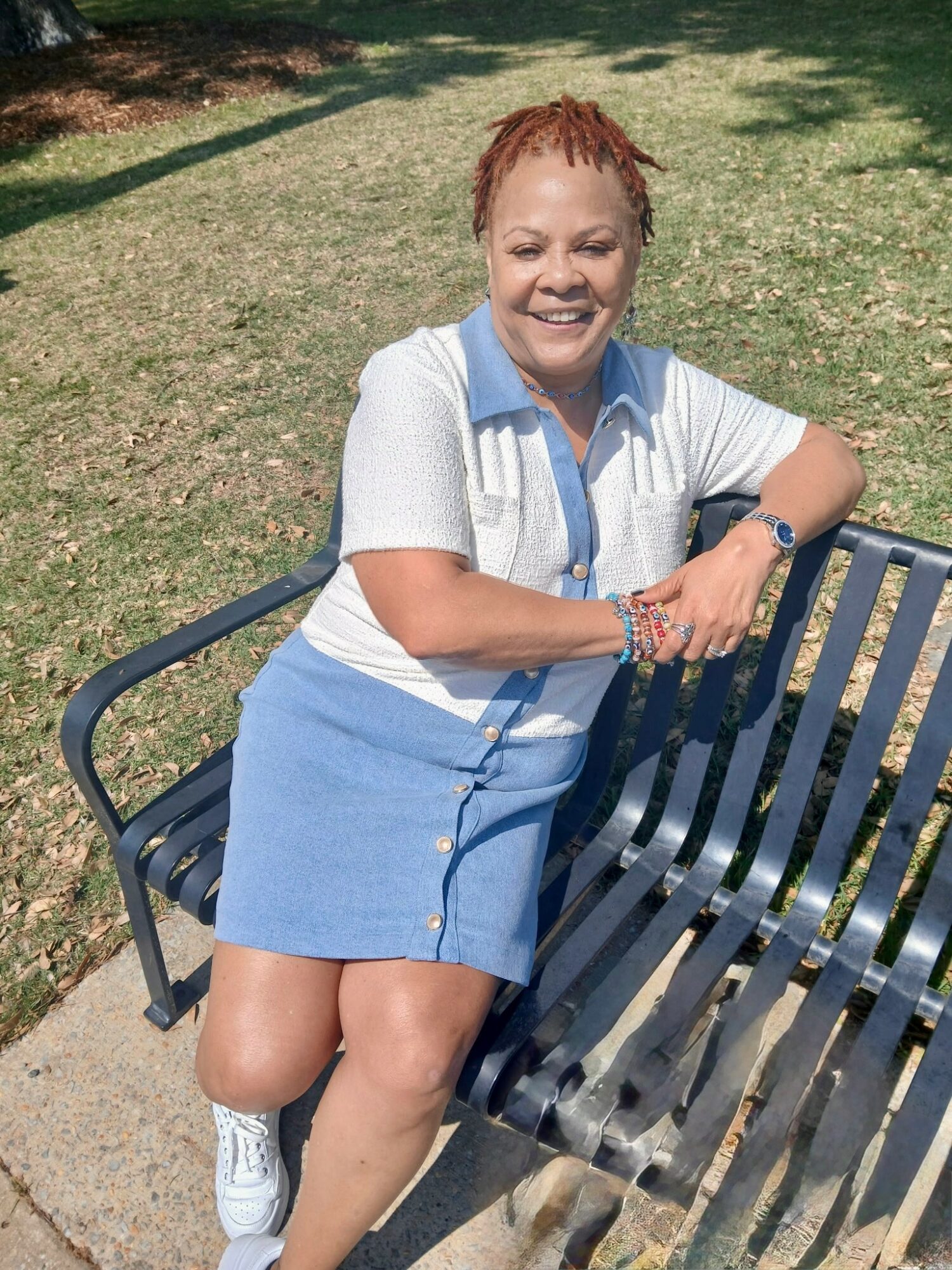 Woman with short hair smiling, leaning on a park bench in sunlight, wearing a striped shirt and blue skirt.