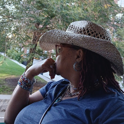 Side profile of a woman wearing a wide-brimmed straw hat, glasses, and jewelry outdoors with trees in background.