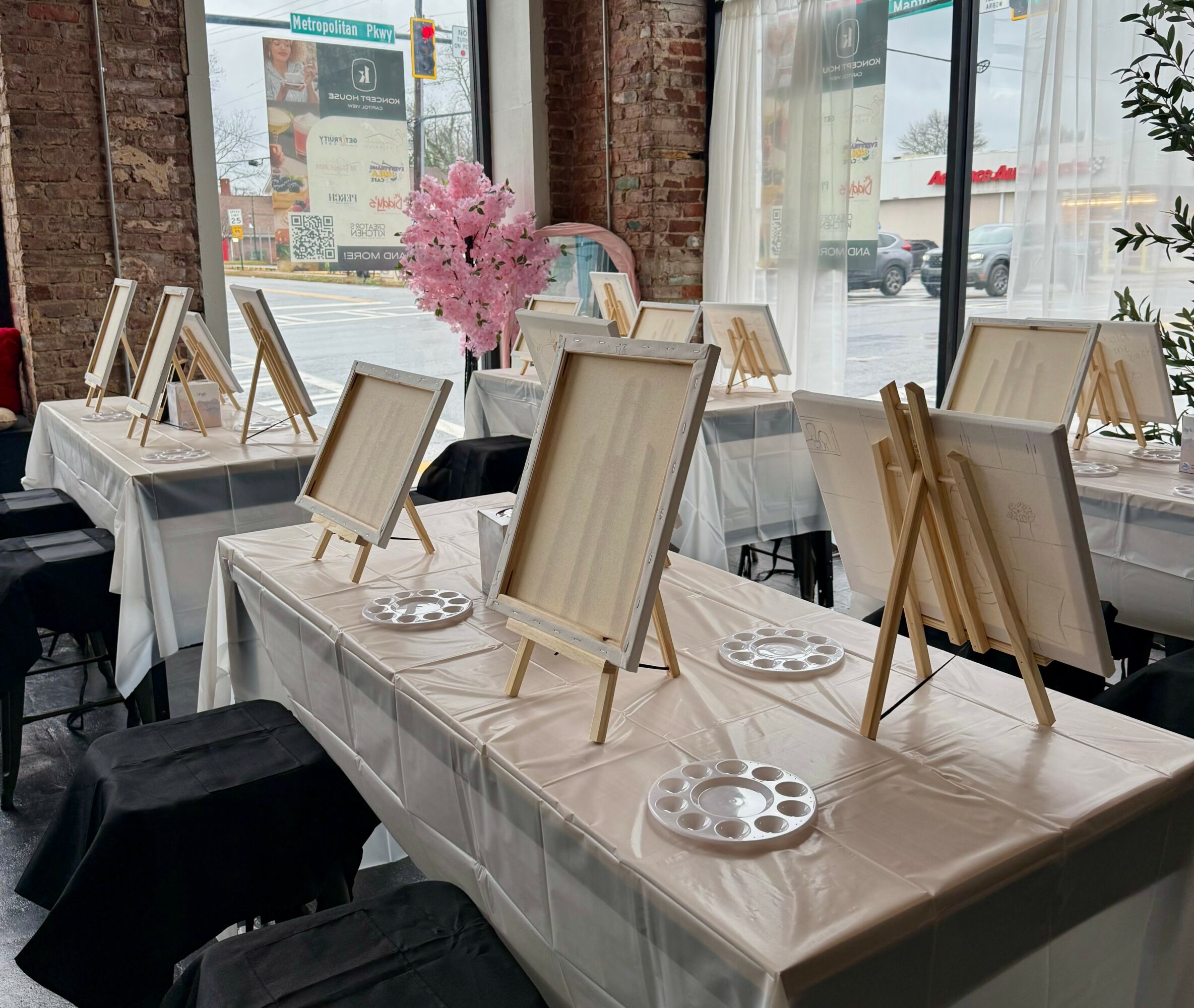 Tables with blank canvases on easels, plates, and a pink floral centerpiece inside a room with large windows.