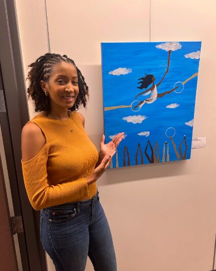 Woman with curly hair in a mustard sweater points at a blue sky painting with a girl on a swing and clouds.