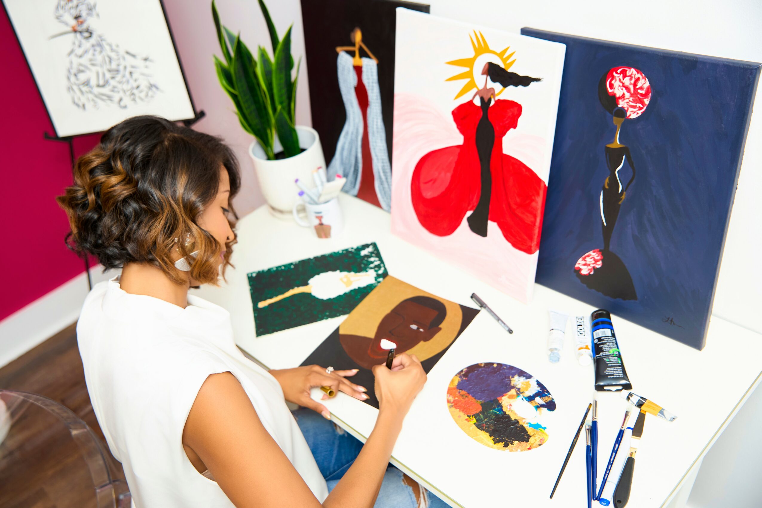 Woman with curly hair painting at a table with colorful abstract and figure artworks, art supplies, and a potted plant nearby.