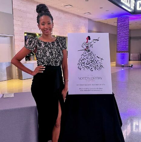 Young woman in black and white dress standing next to a sign on a table in an indoor setting.