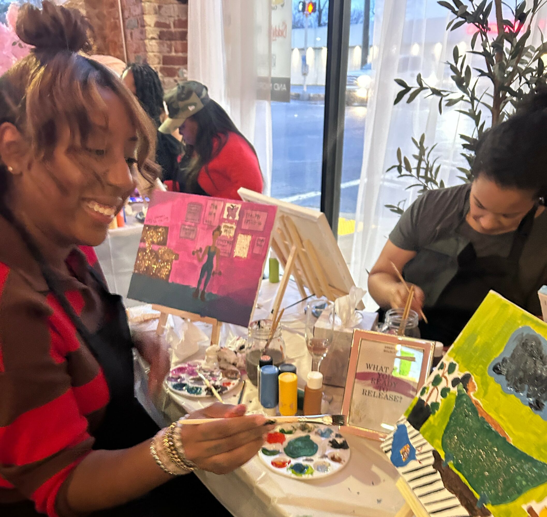 Two women painting at a table with art supplies and colorful paintings, inside a bright cafe with large windows.