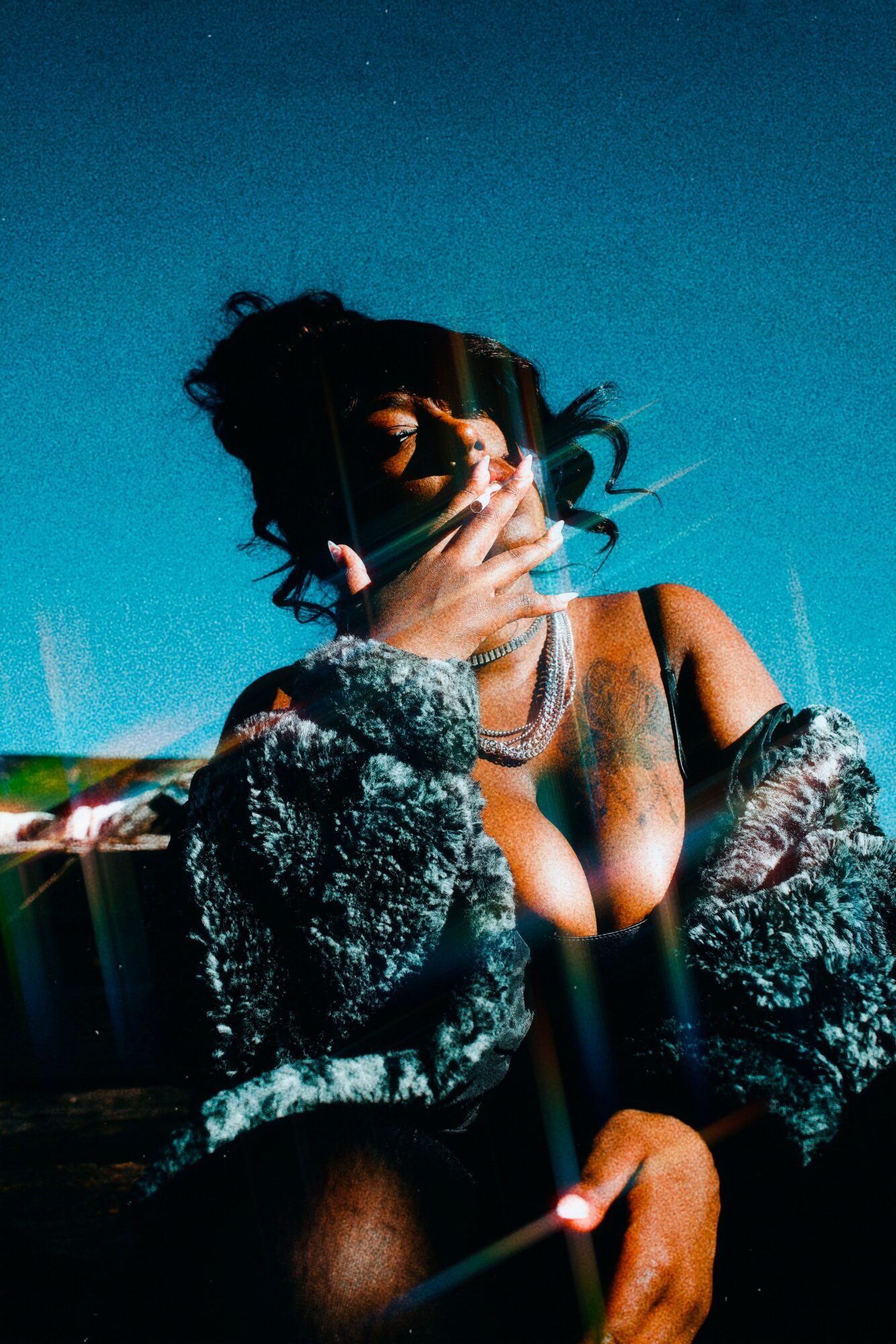 Person with dark hair, wearing a jacket and jewelry, holding a cigarette, against a blue background.