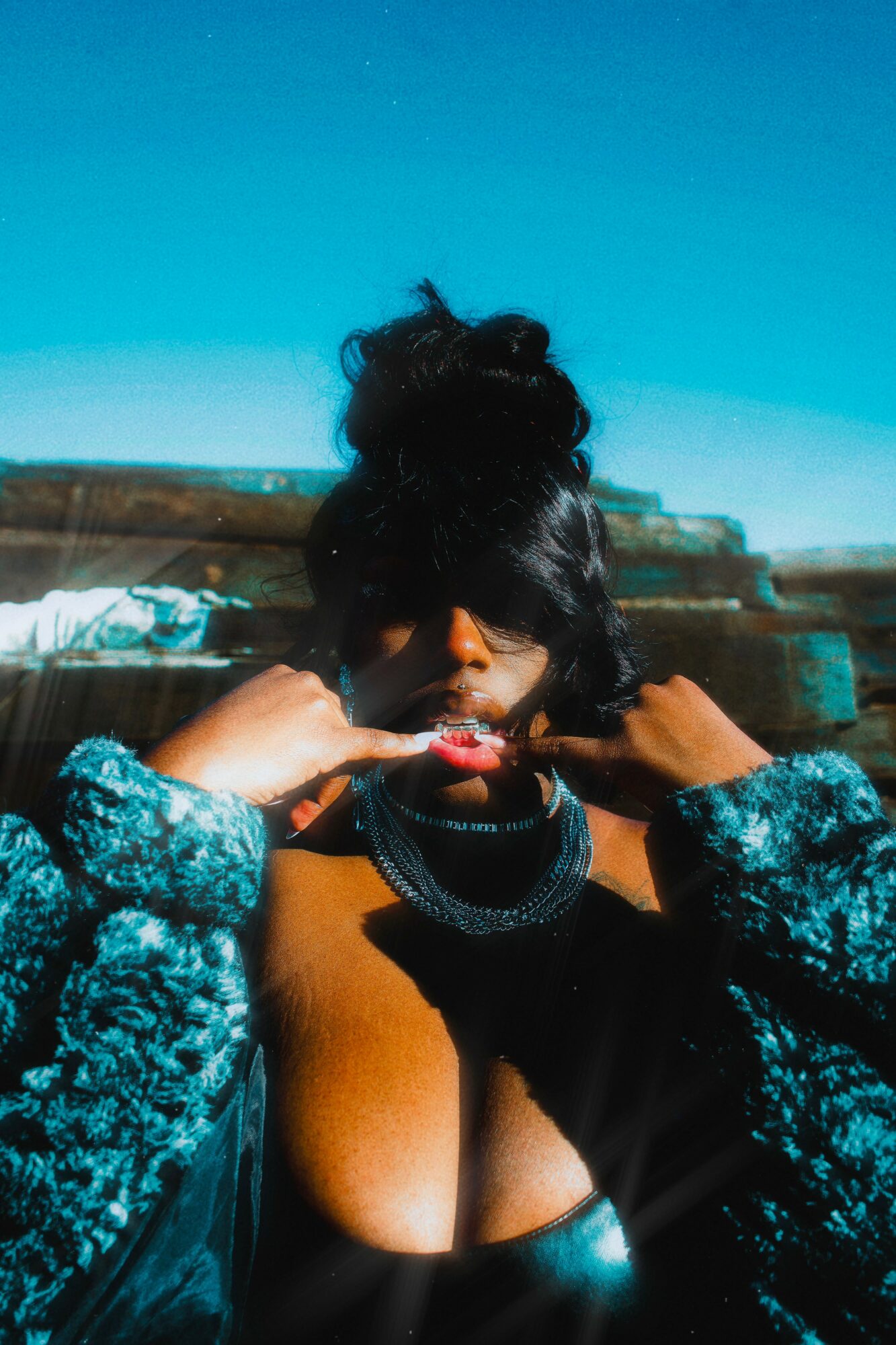 Person with dark hair styled in a bun, wearing a patterned jacket and black top, outdoors with blue sky background.