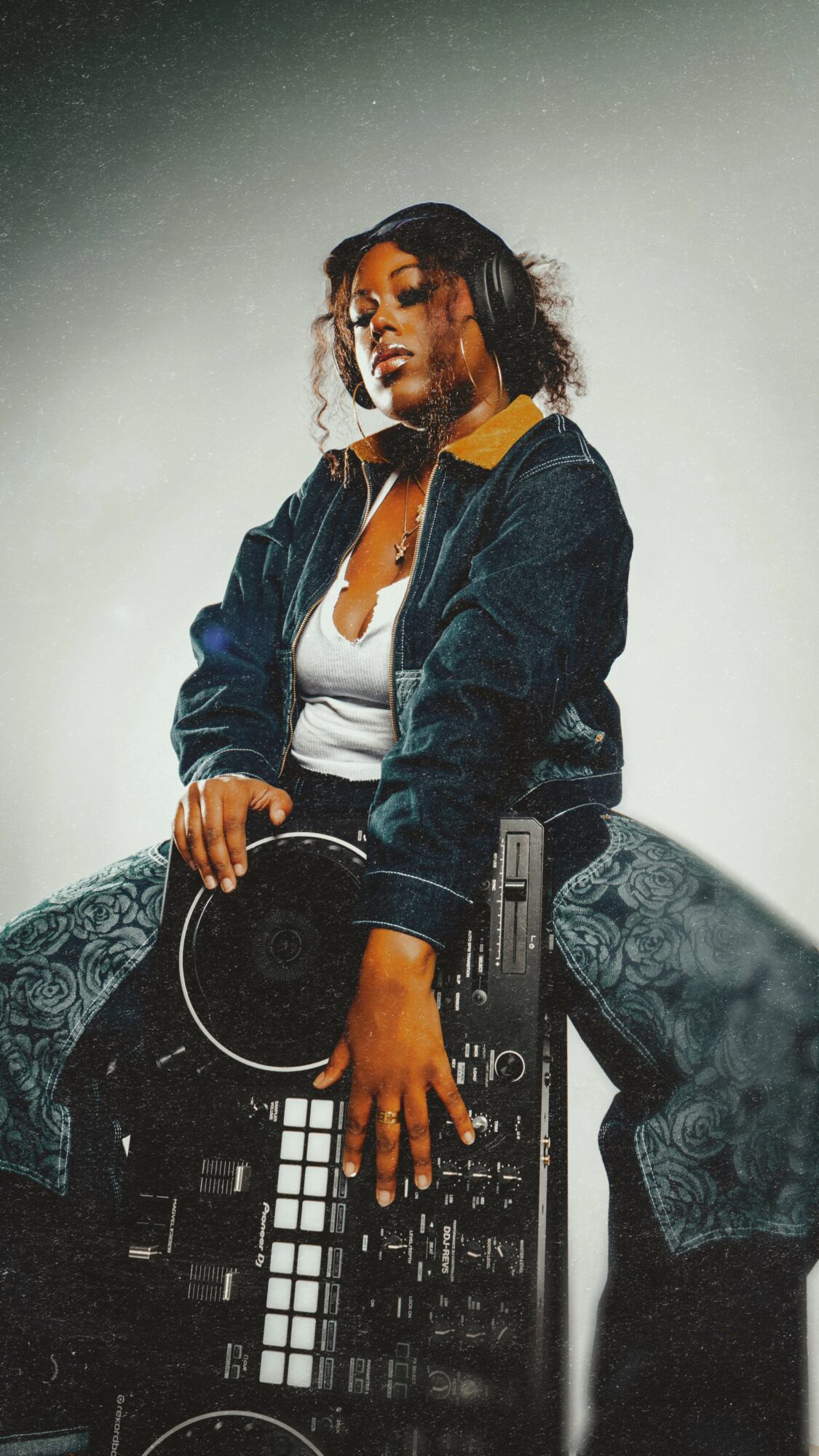 Woman with curly hair wearing a denim jacket and white top, sitting on DJ equipment, looking at camera.