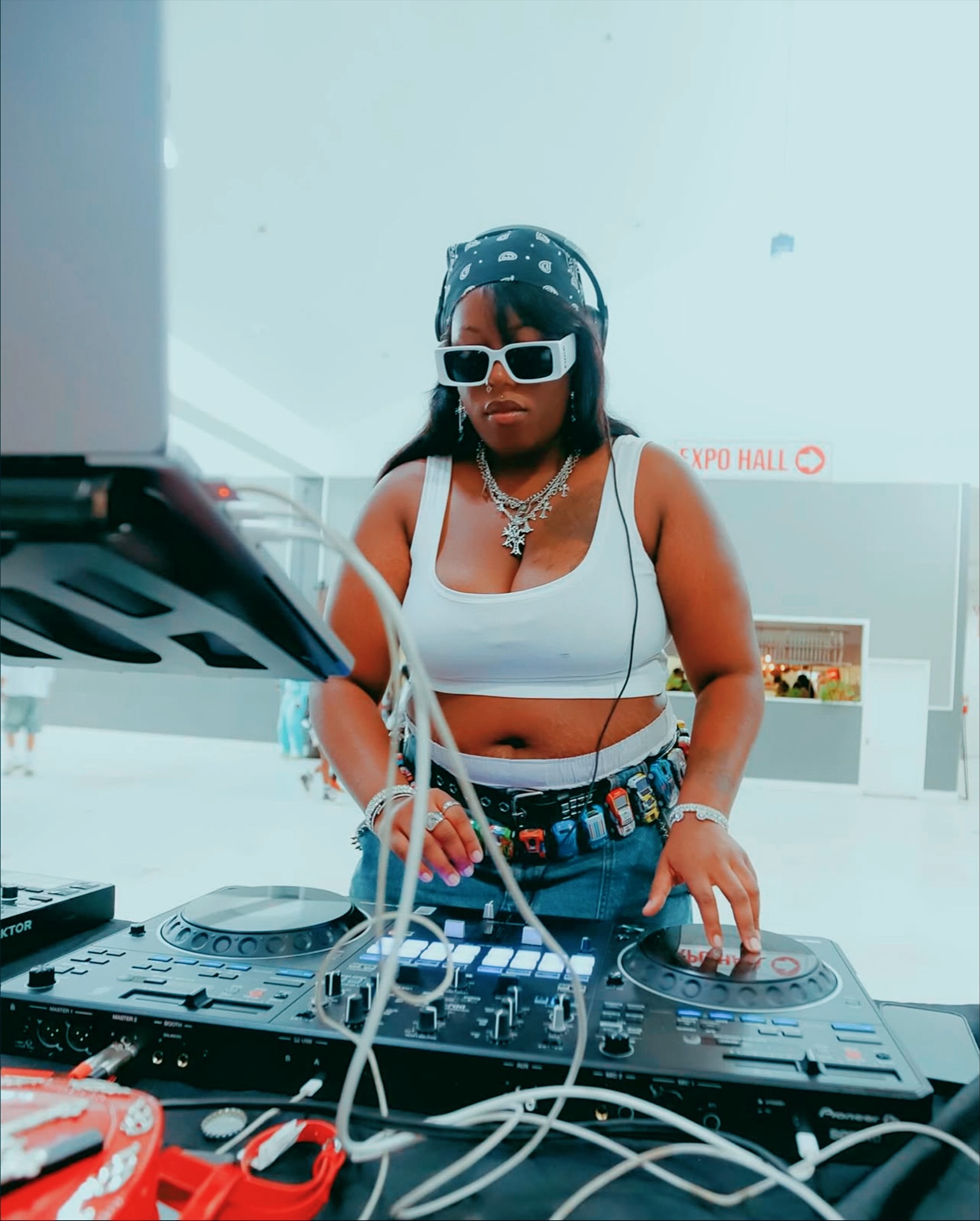 Female DJ wearing sunglasses, headphones, and a white crop top, operating DJ equipment indoors.